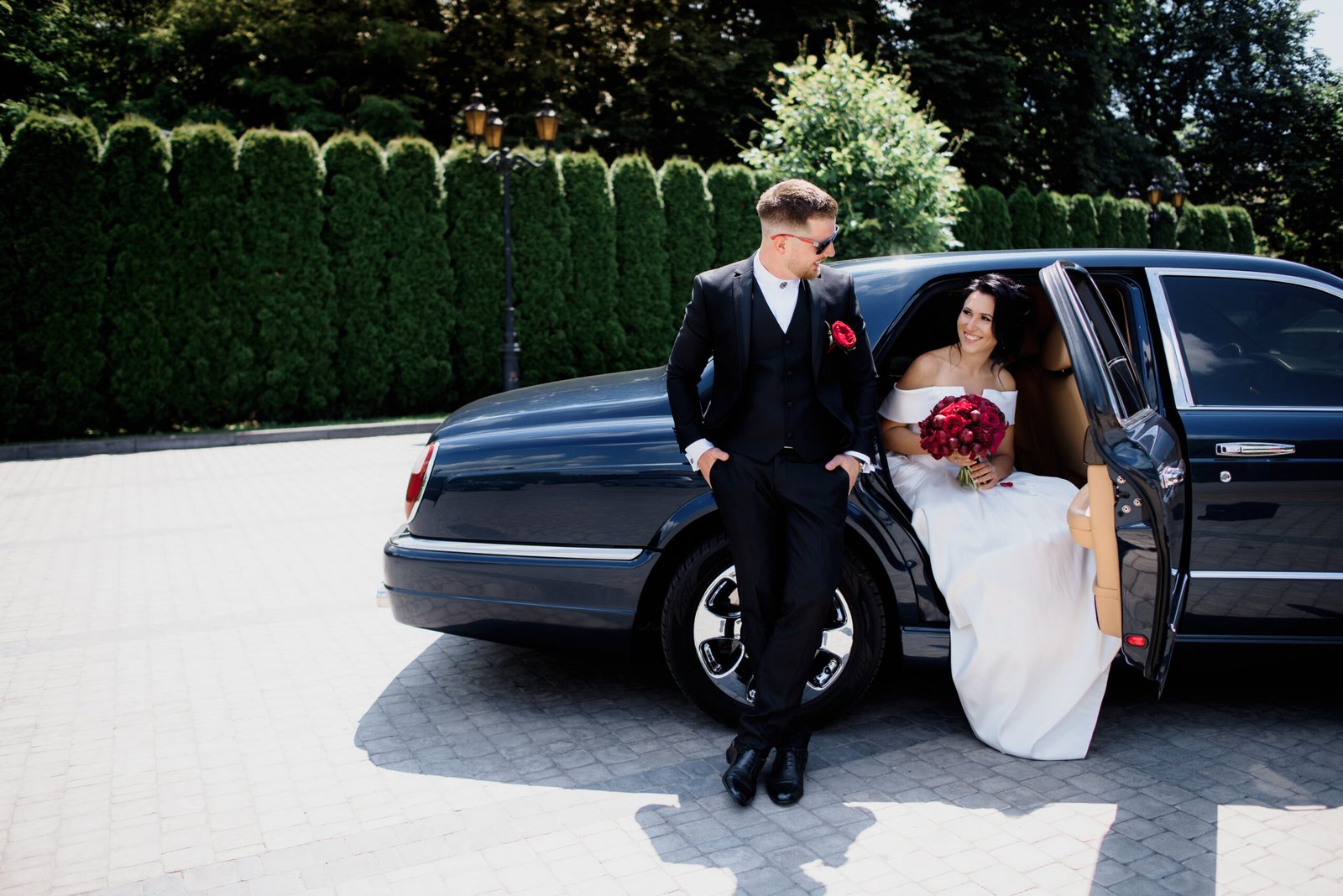 Beautiful wedding couple is smiling in the black car on the sunny day, dressed in elegant wedding outfits with red bouquet